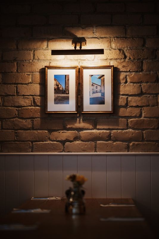 Two framed photographs of Italian streets lit by a picture light on the exposed brick wall at Nucci